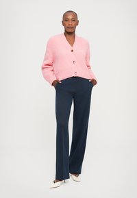 Pink knit cardigan with a V-neck, featuring three large buttons. Paired with navy wide-leg trousers and white heeled shoes.