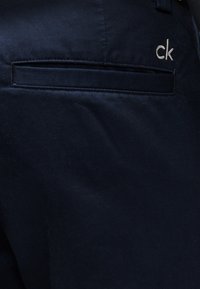 Gros plan sur la poche d'un pantalon en tissu bleu foncé avec une fente horizontale et un petit logo "ck" brodé en blanc au-dessus de la poche.