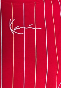 Punainen kangas, jossa on pystysuoria raitoja pastelliväreissä ja näkyvä valkoinen kirjailtu allekirjoitus. Pehmeä rakenne, istuva malli.