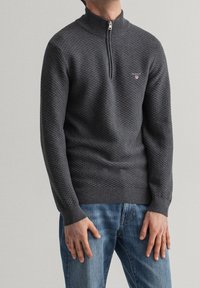 Pull en maille gris foncé avec un col haut et une demi-fermeture éclair, présentant un motif texturé et un petit logo sur la poitrine. Porté avec un jean bleu.