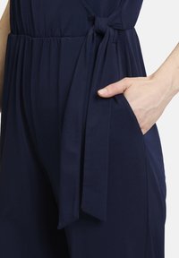 Marineblauwe jumpsuit met een strikceintuur, elastische tailleband en zijzakken. Gladde stof met een relaxte pasvorm en subtiele draperingdetails.