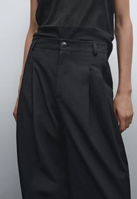 Pantalons noirs taille haute en tissu lisse. Comprend des plis, une fermeture à bouton et une coupe ajustée, avec un design minimaliste.