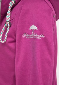 Hanorac fucsia cu glugă, dotat cu un șnur împletit și un logo brodat al unui umbrelu pe mânecă. Textură de bumbac neted.