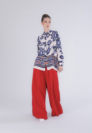 Femme debout portant un pantalon rouge ample, un chemisier blanc avec des motifs floraux et géométriques bleus, et des chaussures blanches pointues sur un fond uni.