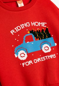 Sudadera roja con un camión pixelado azul y un árbol de Navidad, el texto dice "VOLVIENDO A CASA POR NAVIDAD", detalles en blanco y cuello de canalé.