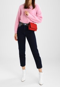 Rosa fluffig tröja, mörka denim jeans, vita ankelstövlar och en röd handväska med scallopkant. Avslappnad outfit med varierande texturer och färger.