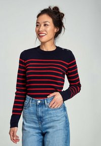 Maglione blu navy con righe orizzontali rosse, scollatura tonda e dettagli a bottoni sulla spalla, abbinato a jeans azzurri a vita alta.
