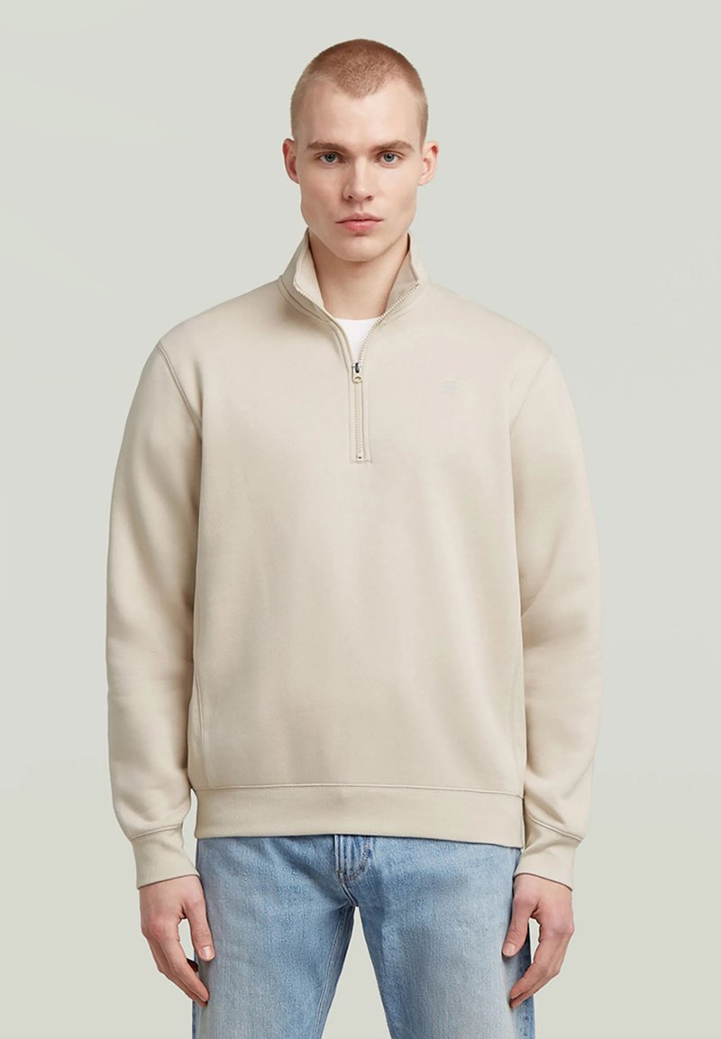 Homme portant un sweat à col montant beige à fermeture éclair partielle et un jean bleu clair, debout devant un fond clair uni.