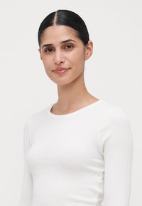 Jeune femme aux cheveux bruns lissés en arrière, portant un haut à manches longues côtelé blanc, souriant doucement contre un fond clair et uni.