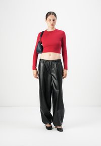Woman wearing a red long-sleeve crop top, black wide-leg leather pants, black heels, and a small black shoulder bag, standing on a white floor.