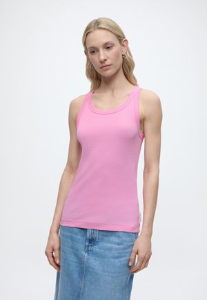 Ženska z dolgimi blond lasmi, oblečena v oprijeto rožnato majico brez rokavov in modro jeansa krilo, na enobarvni belem ozadju.