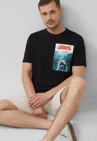 Zwarte katoen t-shirt met een witte en blauwe grafische print van een haai en het woord "JAWS" in vette rode letters.