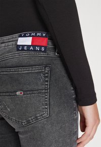 Top negro de manga larga combinado con jeans gris oscuro que presentan un parche visible con el logo de TOMMY JEANS y un pequeño logo en contraste en el bolsillo.