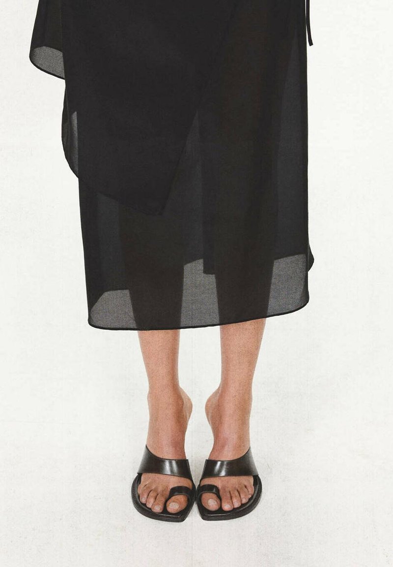 Jambes portant des sandales noires à bout ouvert avec des boucles pour les orteils et une jupe midi noire transparente sur un fond clair.