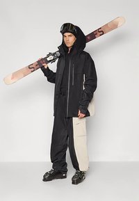 Man in een zwart en beige skikostuum, met een helm en skibril, die een snowboard op zijn schouder houdt, staande tegen een lichte effen achtergrond.