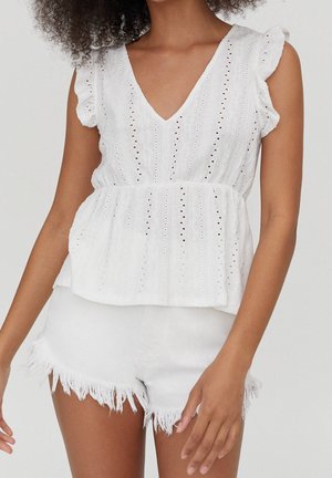 Femme portant un chemisier blanc sans manches en broderie anglaise avec col en V et manches à volants, ainsi qu'un short en jean blanc effiloché, debout devant un fond uni.