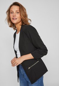 Blazer preto com um corte ajustado, apresentando mangas de três quartos e detalhe de bolso com fecho. Usado sobre uma blusa branca, combinado com jeans azuis.