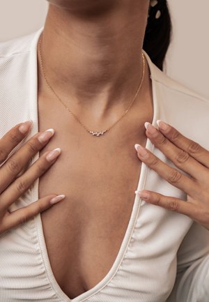 Woman wearing a gold necklace with small diamonds, dressed in white ribbed fabric top with a deep neckline, hands near collarbone showing French manicure.