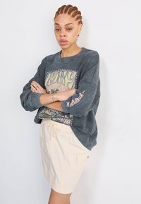 Grå hettegenser med teksturert stoff, med en fargerik grafisk design foran og rullede ermer. Kombinert med lyse beige shorts.
