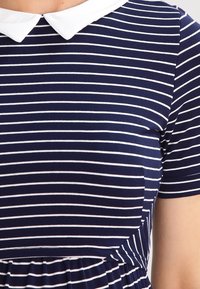 Marinblå klänning med vita horisontella ränder, utrustad med en vit Peter Pan-krage och korta ärmar. Tyget är mjukt och stretchy.