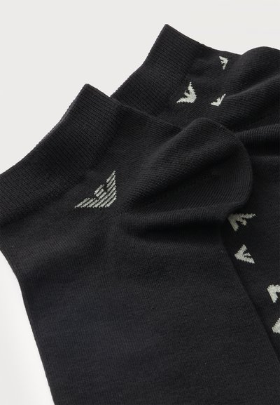 Paire de chaussettes noires chevilles avec un petit logo aile blanche près du bord-côte sur fond uni.