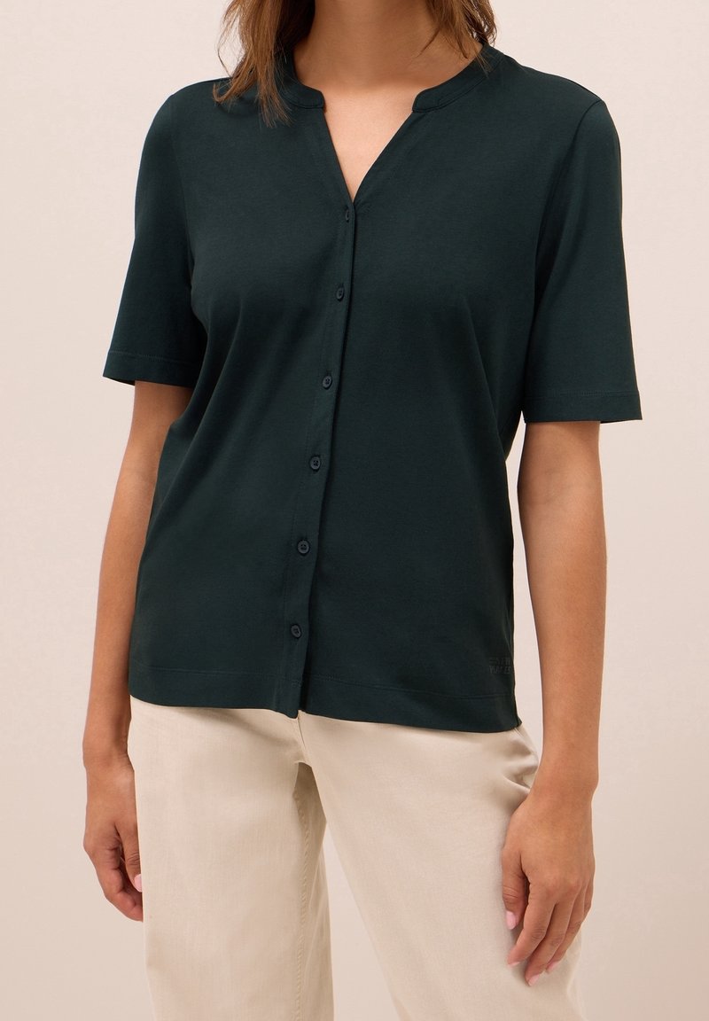 Femme portant une chemise boutonnée à manches courtes vert foncé avec un pantalon beige sur un fond uni.