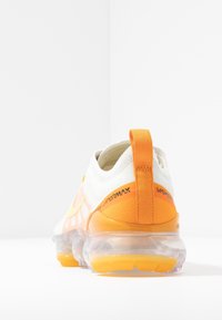 Vit sneaker med orange accenter, med en genomskinlig luftsåle och texturerad överdrag. Bakomdragslapp för enkel påtagning.