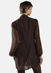 Braunes Kleid mit durchsichtigen langen Ärmeln, tailliertem Oberteil und verdecktem Reißverschluss im Rücken. Glatte Textur mit leicht verlängertem Saum und puffigen Schultern.