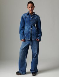 Γυναίκα που φοράει υπερμεγέθη τζιν μπουφάν και τζιν με φαρδιά πόδια, με μαύρα πλατφόρμες με στρας, ποζάροντας μπροστά σε απλό γκρί φόντο.