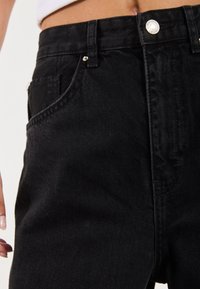 Černé džínové kalhoty s vysokým pasem, předními kapsami a kovovým knoflíkovým zapínáním. Tkanina má hladkou strukturu s jemnými vzory tkaní.