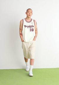 Bela mrežasta majica bez rukava z maroon detalji in besedilom "Originals 33", v kombinaciji z svetlo bež šortami in belimi supergami.