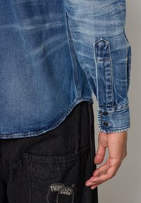 Gros plan d'une personne portant une chemise en jean bleue et un pantalon noir avec un logo graphique sur la poche, main détendue à ses côtés.