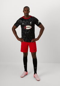 Camiseta de fútbol negra con un sutil patrón geométrico, detalles rojos, pantalones cortos rojos, calcetas negras y botas rosas con logotipos blancos de Nike.