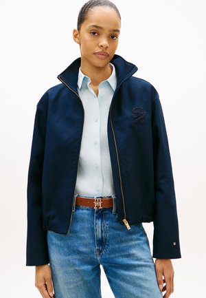Marineblaue Jacke mit Reißverschluss und hohem Kragen, besticktem Logo und elastischem Saum; kombiniert mit hellblauem Hemd und braunem Gürtel, getragen zu geraden Jeans.