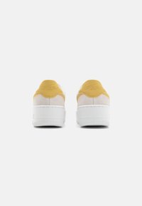 Białe sneakersy z żółtymi akcentami, zamszowa cholewka i gumowa podeszwa o fakturowanej powierzchni. Z tyłu wyeksponowane wypukłe logo.