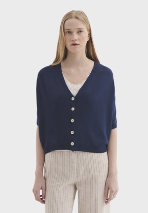 Kvinde med langt blondt hår iført en marineblå knappet cardigan over hvid blondetop og beige bukser med striber, stående mod en ensfarvet baggrund.