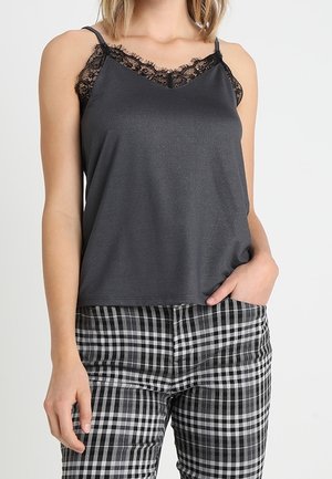 Vrouw draagt een donkergrijs hemdje met zwarte kanten rand en zwart-witte geruite broek, hand nonchalant in de zak.