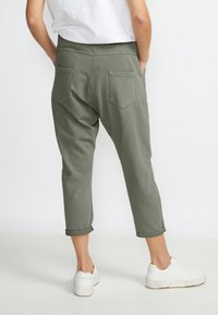 Pantalon cropped vert olive en coton doux. Présente une coupe décontractée, des poches arrière visibles et des ourlets roulés. Porté avec des baskets blanches.