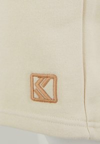 Beige cotton fabric featuring a small, embroidered geometric logo in light brown. Smooth texture with a simple hemline.