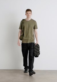 Tricou scurt de culoare verde măslin, pantaloni cargo negri, purtând o geantă din țesătură în dungi și încălțat cu pantofi negri. Geantă crossbody cu curea ajustabilă.