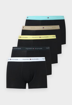 Muskularny mężczyzna stoi boso na jednolitym tle, ubrany w czarne bokserki z żółtym pasem z napisem "Tommy Hilfiger."