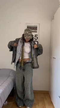 Άτομο που φοράει καπέλο καμουφλάζ, λευκό cropped τοπ, φαρδιά γκρι μπουφάν, φαρδιά γκρι παντελόνια και κίτρινη γραβάτα με σχέδια σε ένα υπνοδωμάτιο.