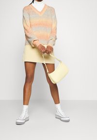 Pull en laine tricoté dans des teintes pastel d'orange et de bleu, assorti à une jupe en velours côtelé crème et un sac à main courbé de couleur jaune clair. Baskets montantes blanches.