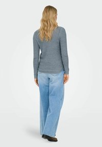Femme aux longs cheveux blonds portant un pull en maille gris et un jean large bleu clair, debout de dos sur un fond uni.