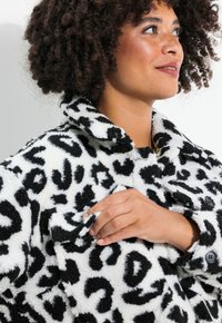 Sort-hvid leopardprint oversized fleecejakke med krave, frontknapper og struktureret stof. Hånd der rører ved jakken.