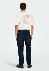Camiseta crema con un estampado gráfico en la parte posterior, combinada con jeans azul oscuro. Presenta un diseño de manga corta y un ajuste relajado.