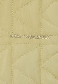 Prošívaná látka v světlé béžové barvě s diamantovým prošíváním. Obsahuje vyšitý název "KARL LAGERFELD" v metalické niti uprostřed.