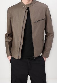 Hnědá lehká bunda na zip s límcem stoje, boční kapsou na zip a hladkou texturou, nošená přes černé tričko a doplněná černými kalhotami.