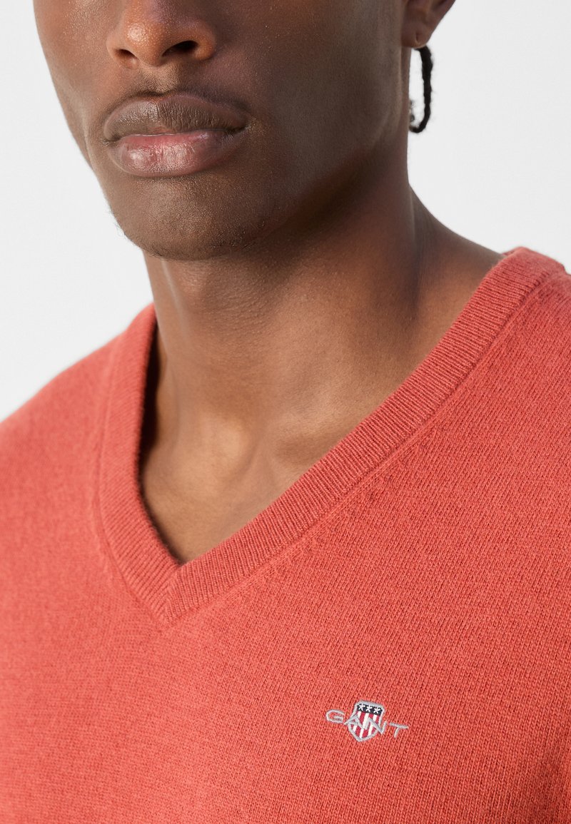 Roter Strickpullover mit V-Ausschnitt, der eine dezente Textur und ein kleines gesticktes Logo auf der Brust aufweist.