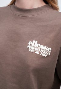 Brūns T-krekls ar baltu "ellesse Perugia Italia Est 1959" logo, kas izdrukāts augšējā krūškurvī tuvumā kakla apakšai.